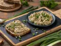 Pyszna pasta z tuńczykiem i jajkiem na ciemnym chlebie, posypana szczypiorkiem. Obok talerz z pastą i świeże warzywa.