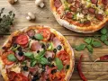 Dwie pizze z dodatkami: salami, pomidory, oliwki, szynka, pieczarki i bazylia.