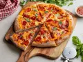 Pikantna pizza diavola z pepperoni i bazylią, pokrojona na kawałki, leży na drewnianej desce. Obok sos i nóż do pizzy.