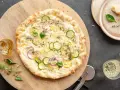 Pyszna pizza bianca z cukinią, cebulą i ziołami, podana na drewnianej desce. Obok buteleczka oliwy i kieliszki z winem.