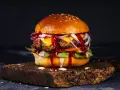 Soczyste burgery z grilla, polane sosem, z serem, cebulą i sałatą, na drewnianej desce.