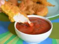 Chrupiący nugget zanurzany w gęstym, czerwonym sosie do nuggetsów. Na talerzu w tle widać więcej złotych kawałków kurczaka.
