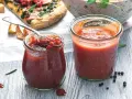 Dwa słoiki domowego sosu pomidorowego, jeden z łyżką pełną aromatycznego sosu. W tle kawałek pizzy i miska z makaronem.