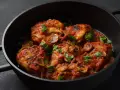 Pyszne udka z kurczaka w bogatym sosie pomidorowym z pieczarkami, posypane świeżą natką pietruszki, podane w czarnej patelni.