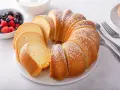 Babka piaskowa przepis babci, puszysta, pokrojona, posypana cukrem pudrem, obok miseczka z owocami i widelce.