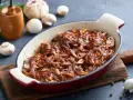 Karkówka pieczona w plastrach z cebulą i sosem, przypominającym ketchup. Danie w naczyniu żaroodpornym, obok grzyby i czosnek.