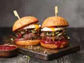 Dwa soczyste burgery z jajkiem sadzonym i grzybami. Idealny stopień wysmażenia burgera podkreśla smak. Obok miseczka sosu.