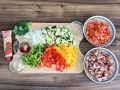 Wszystkie leczo składniki: pokrojone papryki, cukinia, cebula, pomidory, czosnek, kiełbasa, przyprawy i koncentrat pomidorowy.