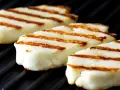 Trzy kawałki halloumi z grilla, z charakterystycznymi brązowymi paskami, leżą na czarnej, grillowej powierzchni.