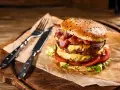Podwójny burger z soczystym mięsem na burgery, serem, bekonem i warzywami, gotowy do zjedzenia z widelcem i nożem.