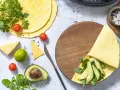 Keto tortilla z awokado i rukolą na drewnianej desce, obok pomidorki, ser i limonka.