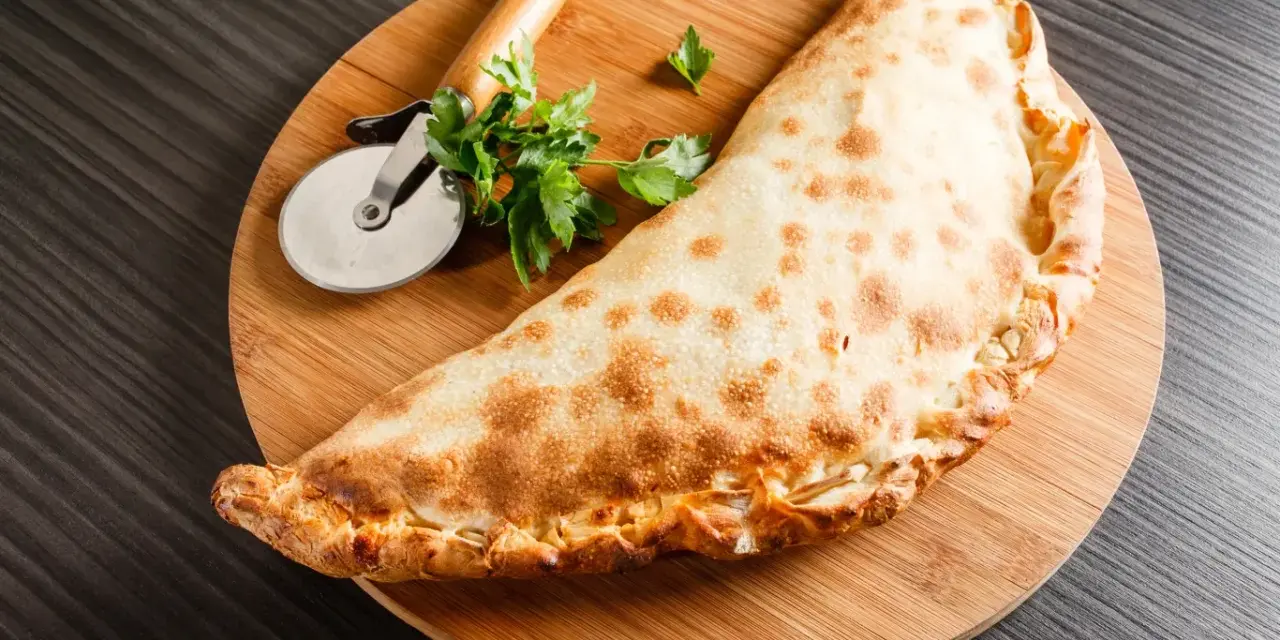 Złocisty calzone, czyli włoska pierożkowa pizza, leży na desce z krążkiem do pizzy i natką pietruszki.