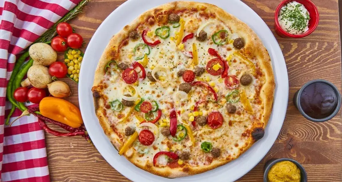 Pizza z mięsem mielonym, pomidorkami, papryką i kukurydzą na białym talerzu, obok sosy i warzywa.