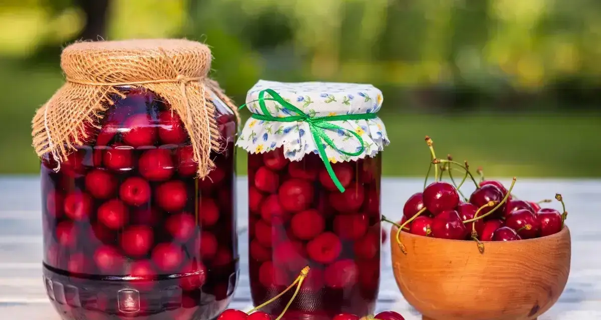 Dwa słoje domowego kompotu z czereśni, jeden z lnianym, drugi z kwiecistym nakryciem, obok miseczka świeżych owoców.