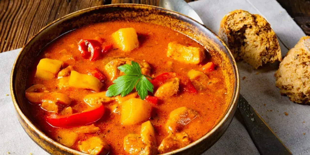 A miseczka aromatycznej zupy gulaszowej, jak z przepisu babci, z kawałkami mięsa, ziemniaków i papryki, udekorowana natką pietruszki. Obok kromki chleba.