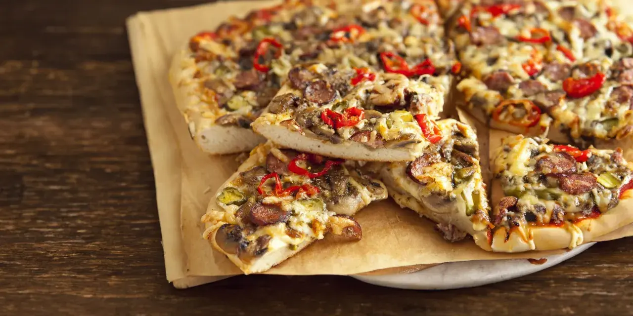 Grube ciasto na pizze z kiełbasą, pieczarkami i papryką, podane na papierze do pieczenia.
