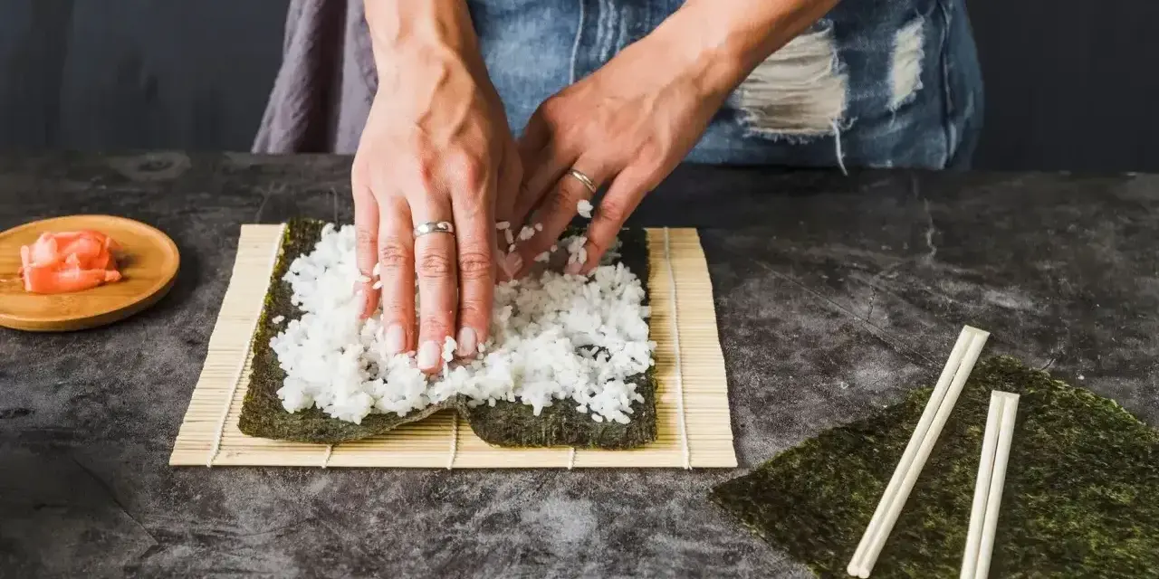 Ręce rozkładają ryż na nori, przygotowując zaprawę do sushi na 500g ryżu. Obok leżą pałeczki i imbir.