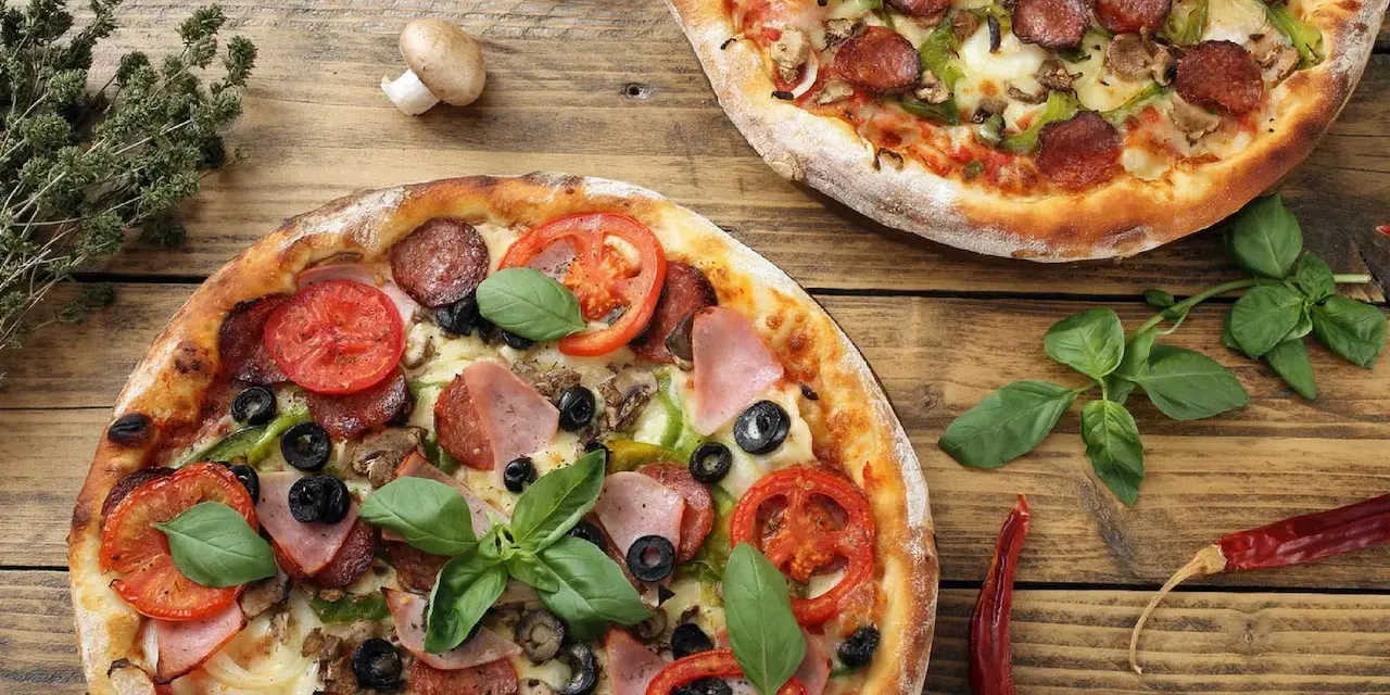 Dwie pizze z dodatkami: salami, pomidory, oliwki, szynka, pieczarki i bazylia.
