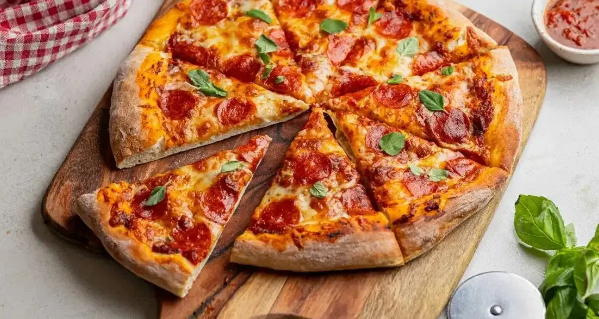 Pikantna pizza diavola z pepperoni i bazylią, pokrojona na kawałki, leży na drewnianej desce. Obok sos i nóż do pizzy.