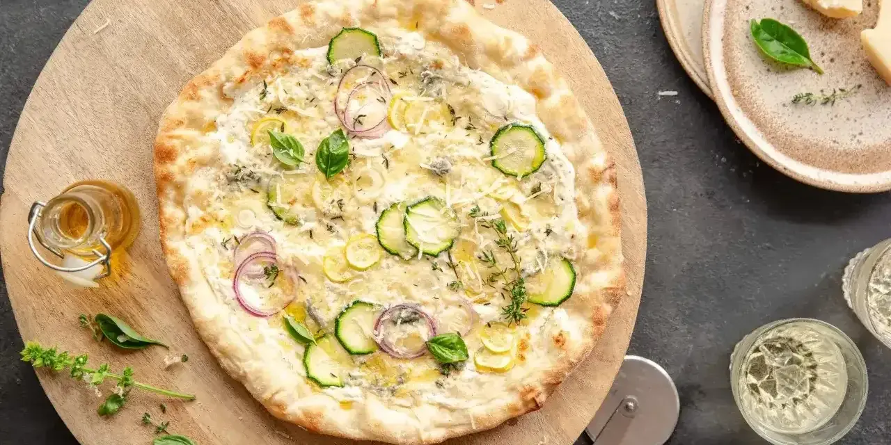 Pyszna pizza bianca z cukinią, cebulą i ziołami, podana na drewnianej desce. Obok buteleczka oliwy i kieliszki z winem.