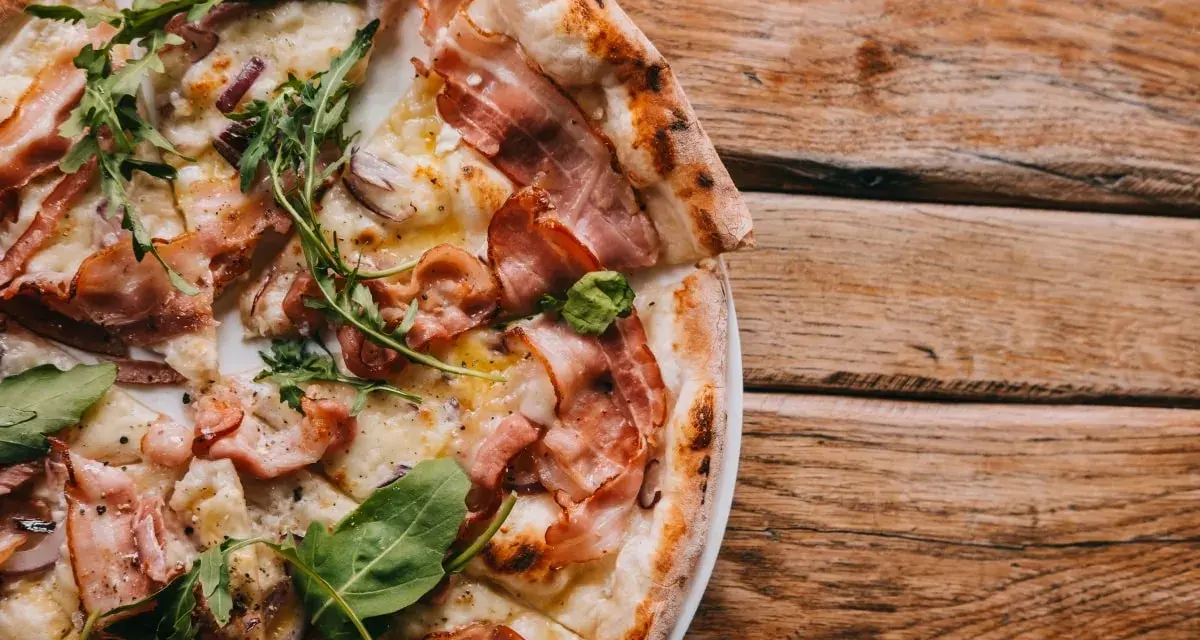 Pyszna pizza carbonara z chrupiącym boczkiem i rukolą, podana na drewnianym blacie.