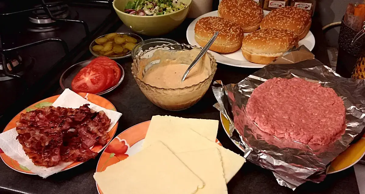 Składniki do przygotowania idealnego cheeseburgera: bułki, surowe mięso, ser, bekon, pomidory, ogórki i sos.