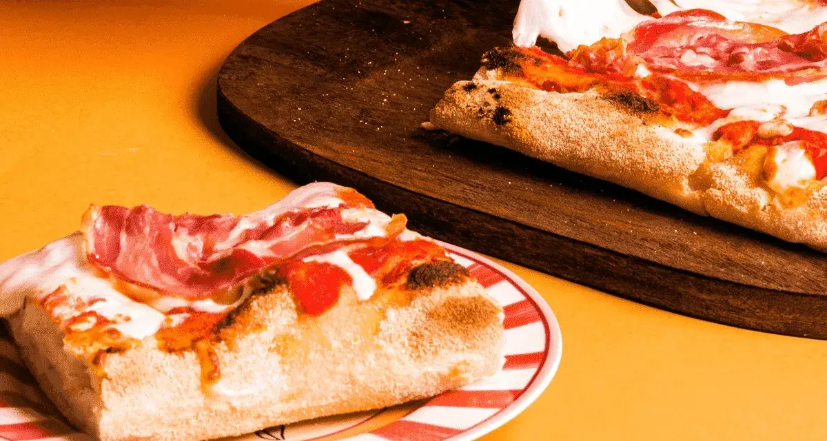 Kawałek pysznej pizzy rzymskiej z szynką i ciągnącym się serem na talerzu, obok reszta na desce.