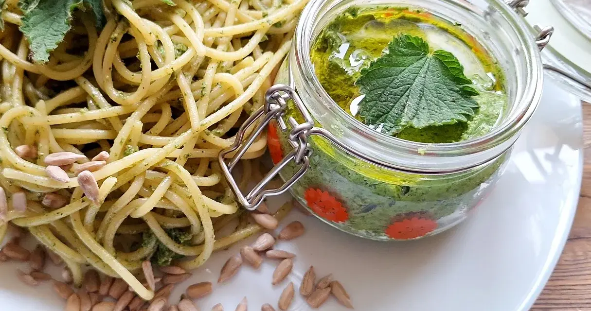 Makaron z pesto z pokrzywy, ozdobiony świeżymi liśćmi pokrzywy i pestkami słonecznika. Obok słoiczek z domowym pesto.