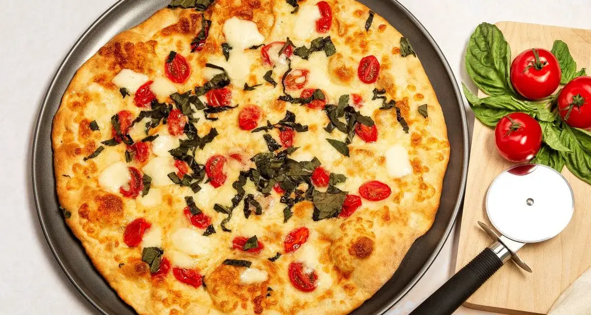 Pyszna pizza z serka wiejskiego, pomidorkami koktajlowymi i bazylią. Obok leży krajalnica do pizzy i świeże pomidory.