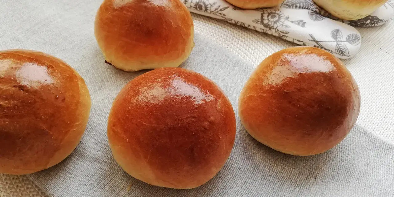 Pięć puszystych bułek maślanych, lśniących od pieczenia, ułożonych na lnianej serwetce.
