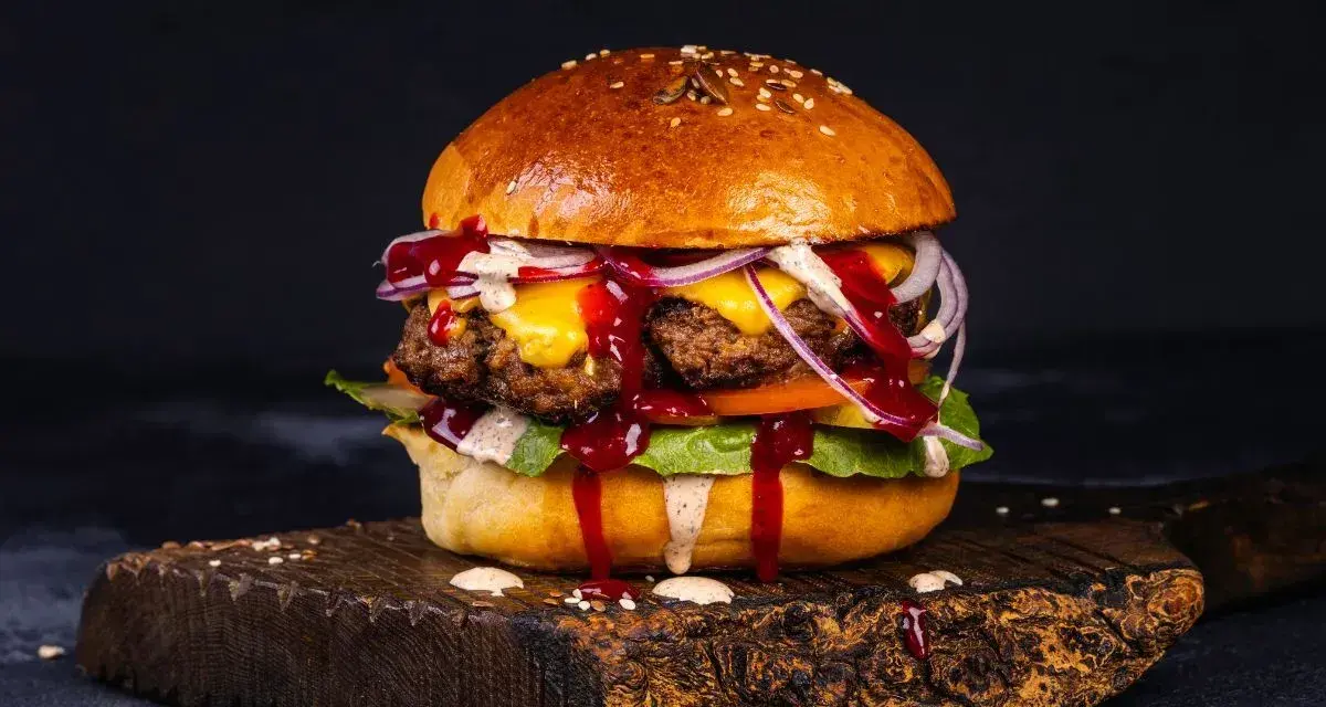 Soczyste burgery z grilla, polane sosem, z serem, cebulą i sałatą, na drewnianej desce.