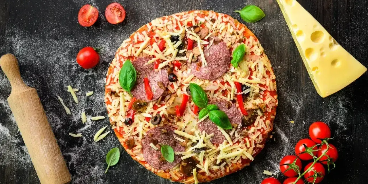 Gotowa pizza z salami, serem i warzywami, obok wałek, pomidorki i ser. Idealna na wieczór z przyjaciółmi, gdy brakuje czasu na przygotowanie ciasta z mąki na pizzę.