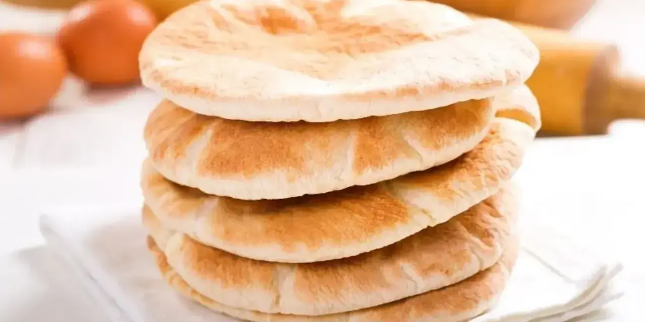 Stos świeżo upieczonego chleba arabskiego na białej serwetce, obok jajka i wałka do ciasta.