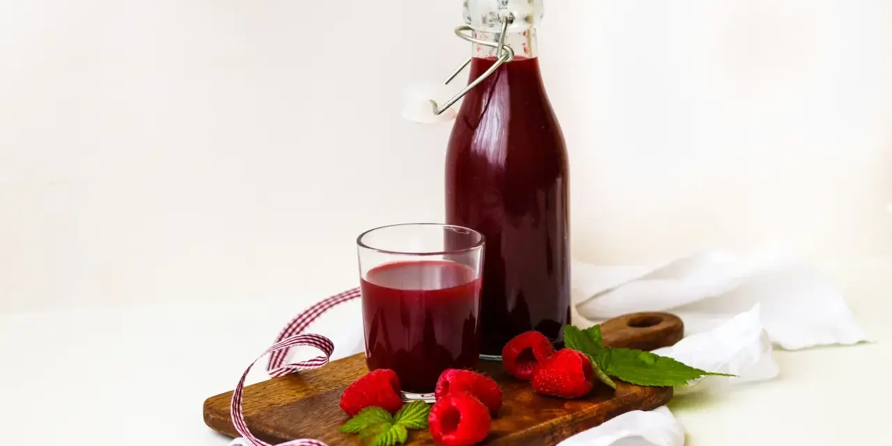 Szklanka i butelka z ciemnoczerwonym sokiem z malin, na drewnianej desce z malinami i listkami mięty.