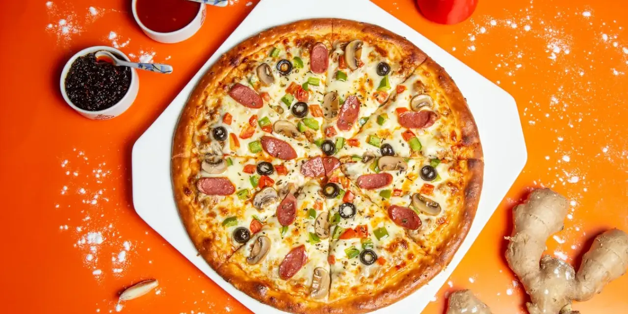 Pizza z kiełbasą, pieczarkami i oliwkami na pomarańczowym tle, obok sosy i imbir.