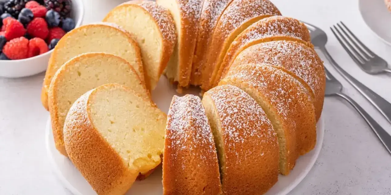 Babka piaskowa przepis babci, puszysta, pokrojona, posypana cukrem pudrem, obok miseczka z owocami i widelce.