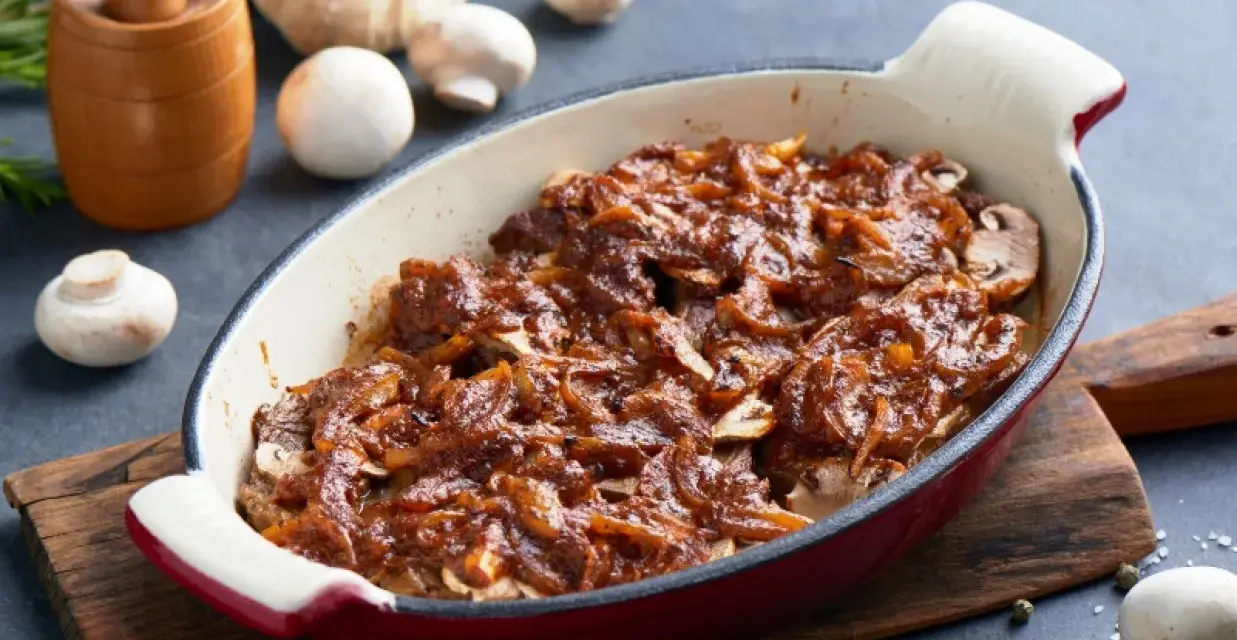 Karkówka pieczona w plastrach z cebulą i sosem, przypominającym ketchup. Danie w naczyniu żaroodpornym, obok grzyby i czosnek.