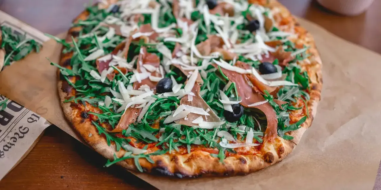 Apetyczna pizza prosciutto z rukolą, oliwkami i parmezanem, podana na papierze.