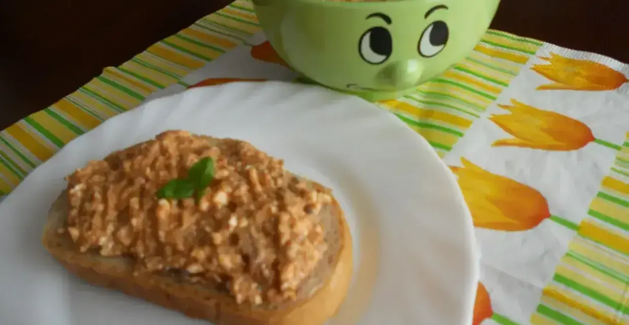 Pyszna pasta z makreli i twarogu na kromce chleba, udekorowana listkiem bazylii. Obok miseczka z pastą i wesołą buźką.