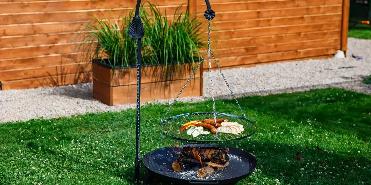 Grill trójnóg z paleniskiem i rusztem, na którym znajdują się kiełbaski, pieczywo i warzywa.