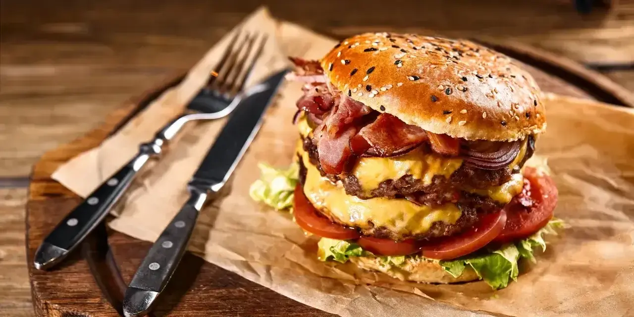 Podwójny burger z soczystym mięsem na burgery, serem, bekonem i warzywami, gotowy do zjedzenia z widelcem i nożem.