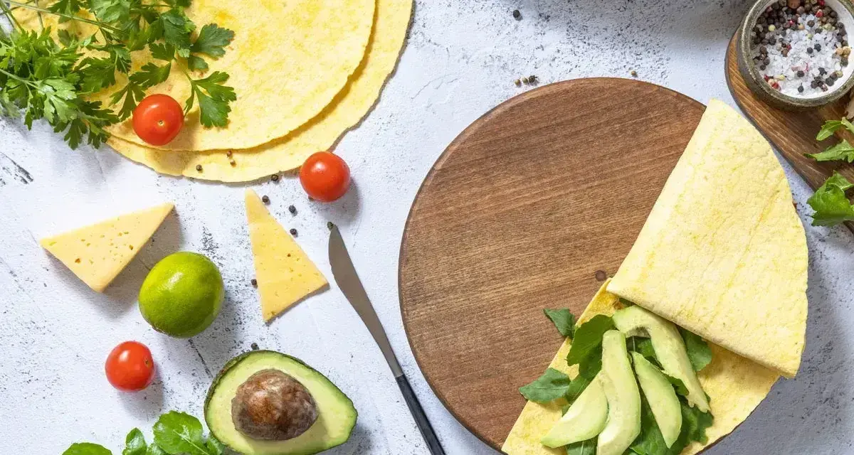 Keto tortilla z awokado i rukolą na drewnianej desce, obok pomidorki, ser i limonka.