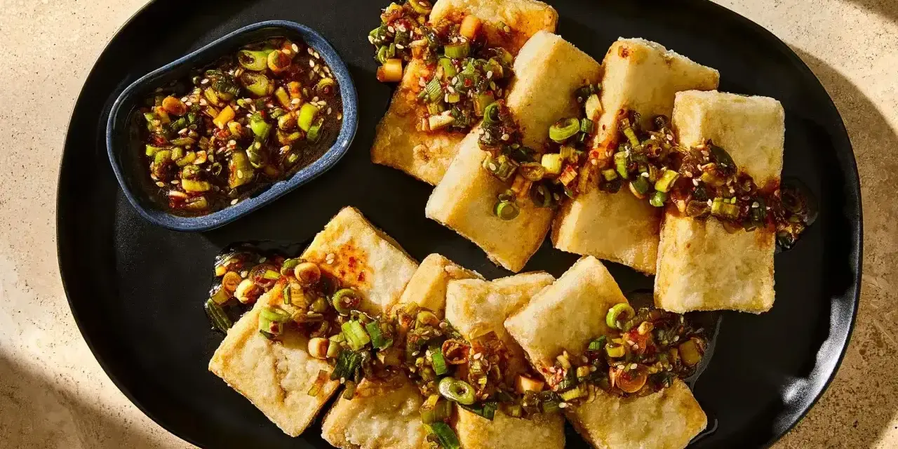 Chrupiące kawałki tofu polane aromatyczną marynatą z dymką i sezamem, podane na czarnym talerzu.