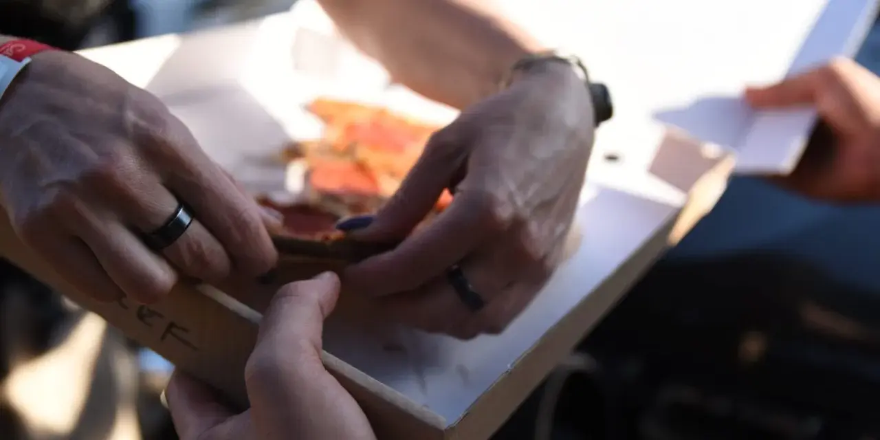 Dwie osoby dzielą się pizzą z pudełka. Właśnie pokazują sobie, jak jeść pizzę, by każdy dostał swój kawałek.