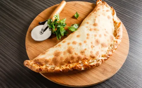 Złocisty calzone, czyli włoska pierożkowa pizza, leży na desce z krążkiem do pizzy i natką pietruszki.