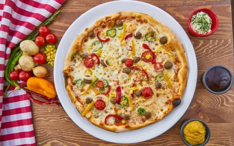 Pizza z mięsem mielonym, pomidorkami, papryką i kukurydzą na białym talerzu, obok sosy i warzywa.