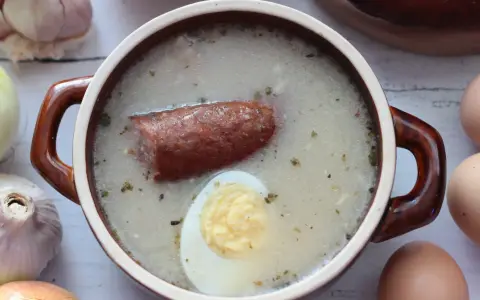 Gęsty, biały żurek z kiełbasą i jajkiem, przygotowany na bazie zakwasu na żurek. Obok czosnek, cebula i jajka.
