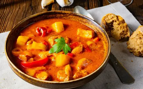 A miseczka aromatycznej zupy gulaszowej, jak z przepisu babci, z kawałkami mięsa, ziemniaków i papryki, udekorowana natką pietruszki. Obok kromki chleba.