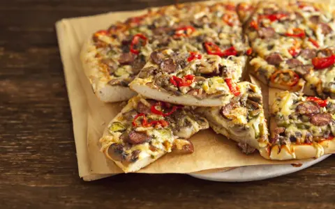 Grube ciasto na pizze z kiełbasą, pieczarkami i papryką, podane na papierze do pieczenia.