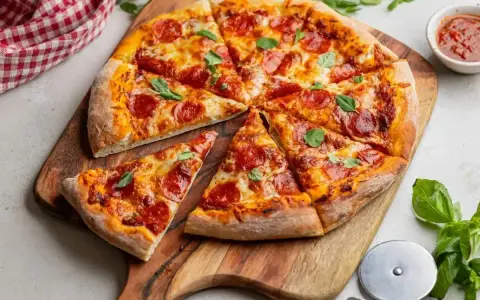 Pikantna pizza diavola z pepperoni i bazylią, pokrojona na kawałki, leży na drewnianej desce. Obok sos i nóż do pizzy.